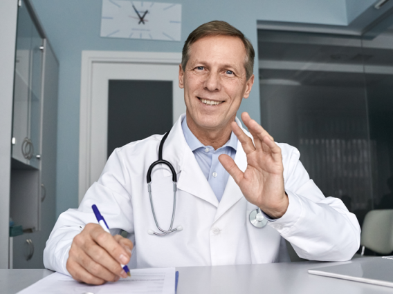 Male doctor sitting at desk looking ahead and waving.